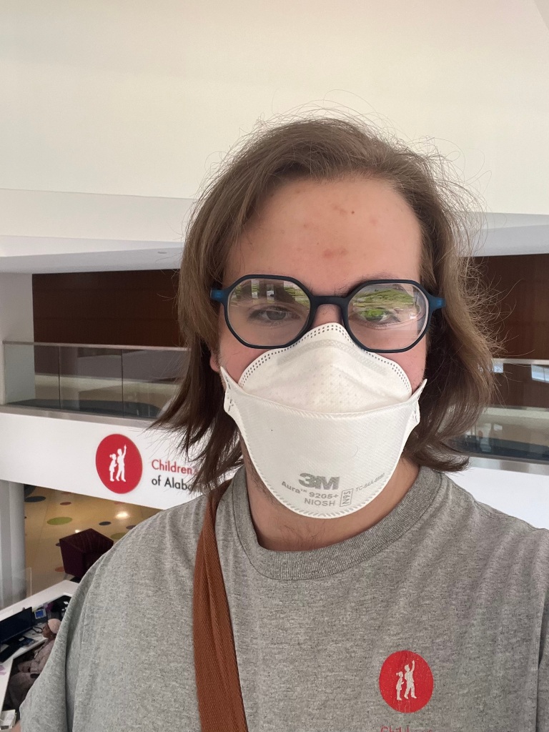 Alternative text: "Derek stands in the atrium of Children's of Alabama. They are wearing a grey shirt with the Children's of Alabama logo, blue glasses, and a mask. They have a light brown strap on their shoulder."