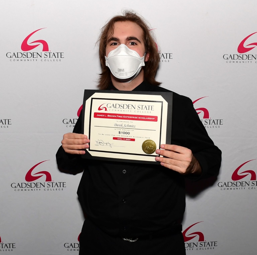 "Derek stands in front of a Gadsden State Community College backdrop wearing all black and a mask. They are holding a certificate with both hands and smiling."